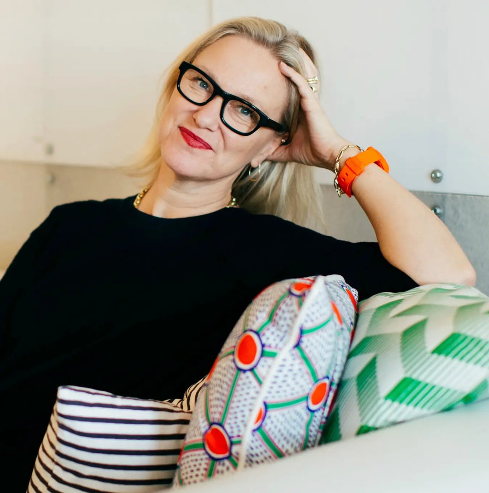 Ghislaine Vinas with glasses and an orange watch sitting on a couch with patterned cushions.