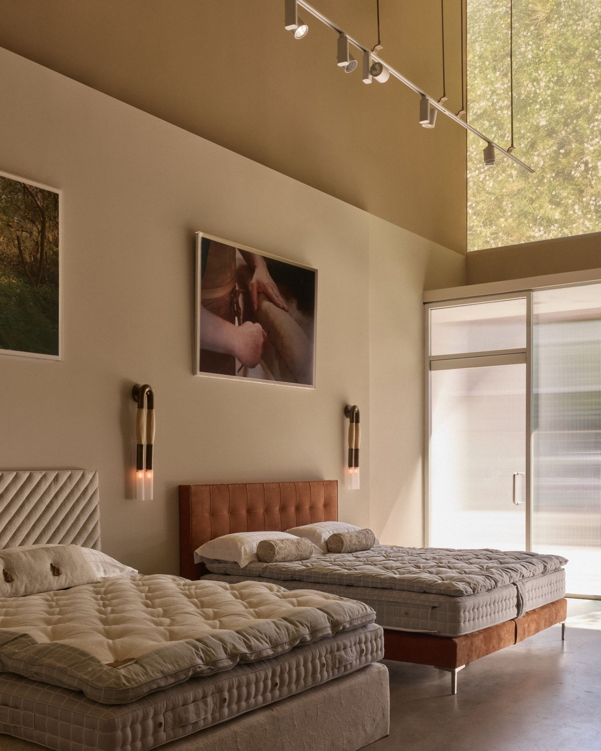 The LA Savoir showroom featuring two beds with quilted beige bedding, a tufted brown headboard, wall-mounted sconces, framed wall art, and a large window with blinds.