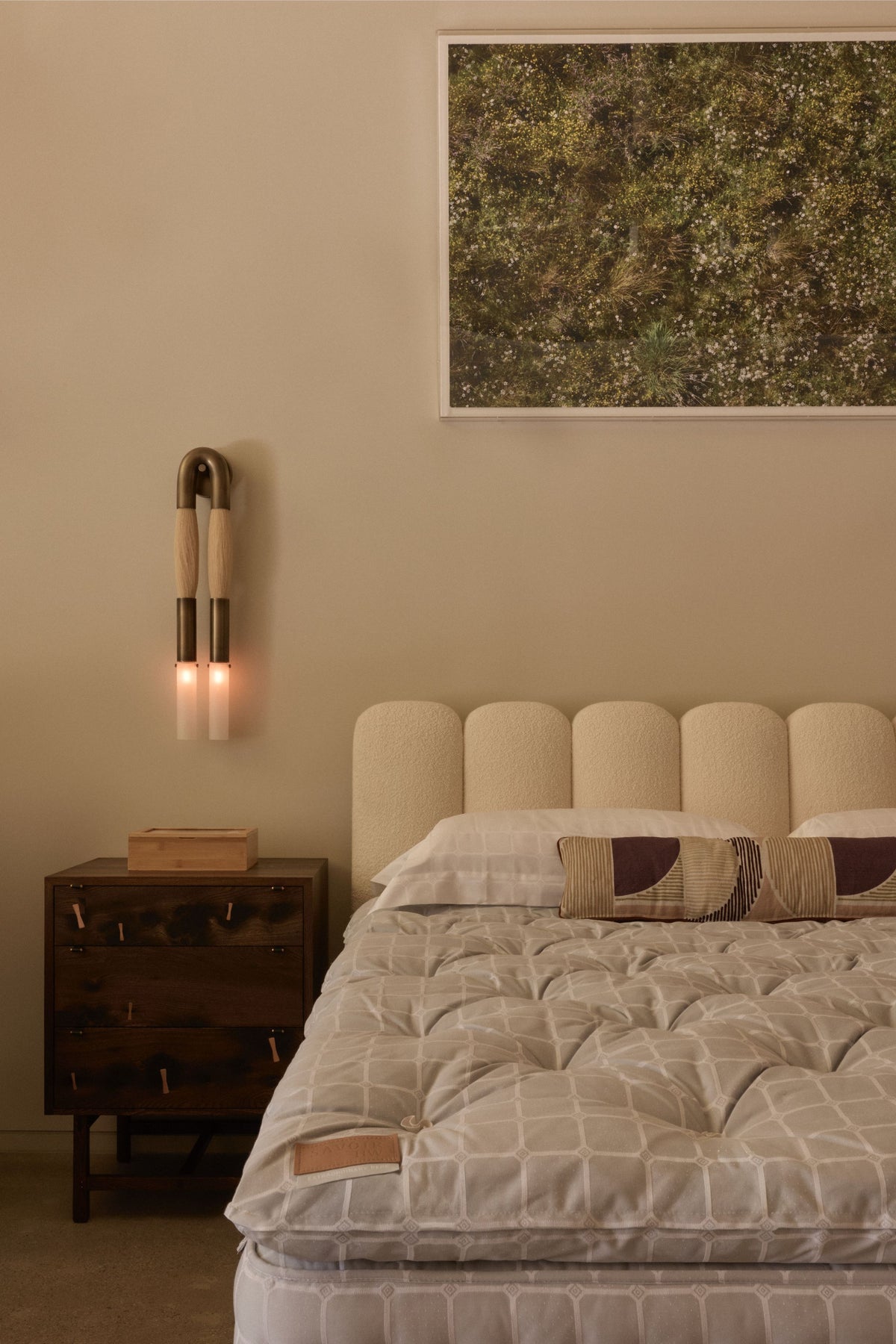The Savoir Los Angeles showroom  featuring the Melrose bed with a cream tufted headboard and patterned quilt, a dark wood nightstand with a wall-mounted sconce, and a framed abstract painting on the beige wall.