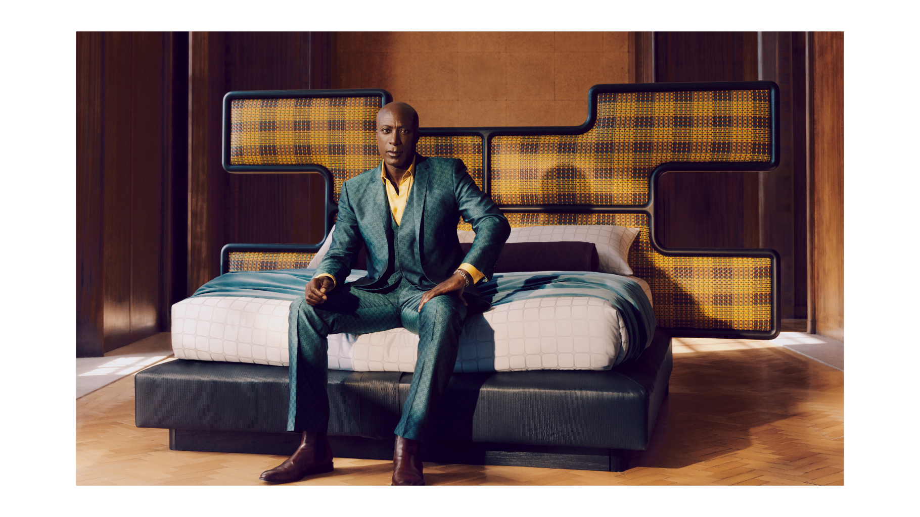 Ozwald Boateng in a suit sitting on a modern bed with kente fabric  headboard.