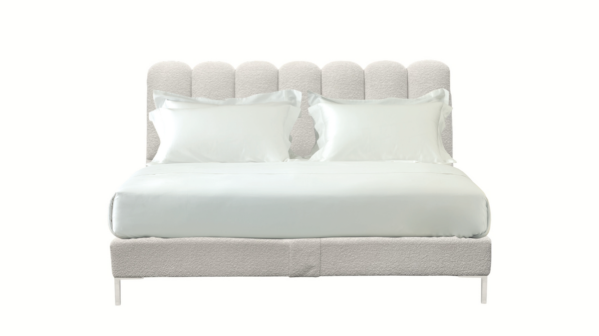 A modern upholstered bed with a tall, scalloped headboard and matching base in a light beige textured fabric, styled with white bedding and two white pillows against a solid white background.