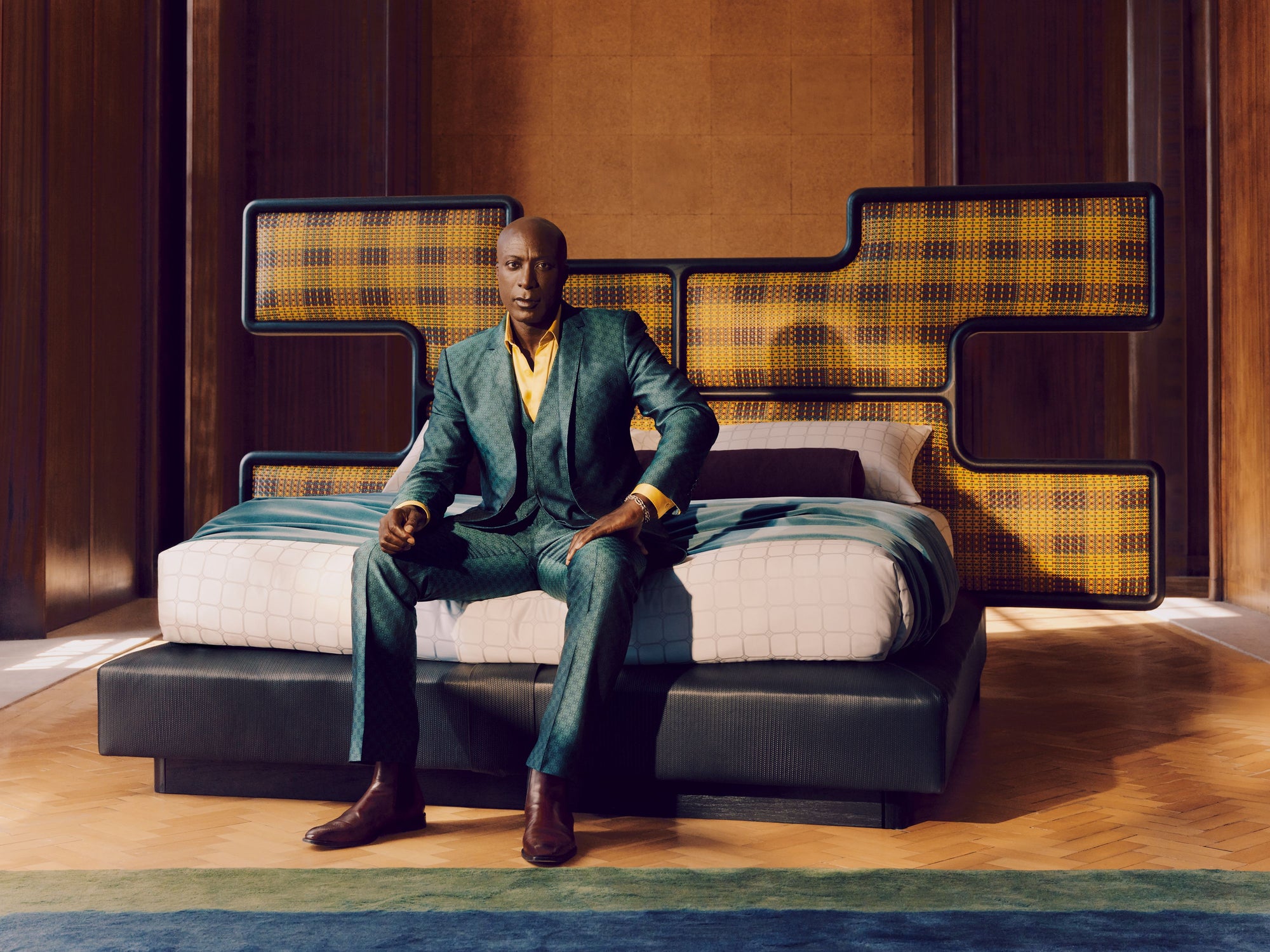 Ozwald Boateng in a suit sitting on a modern sofa with a patterned headboard in a stylish room.