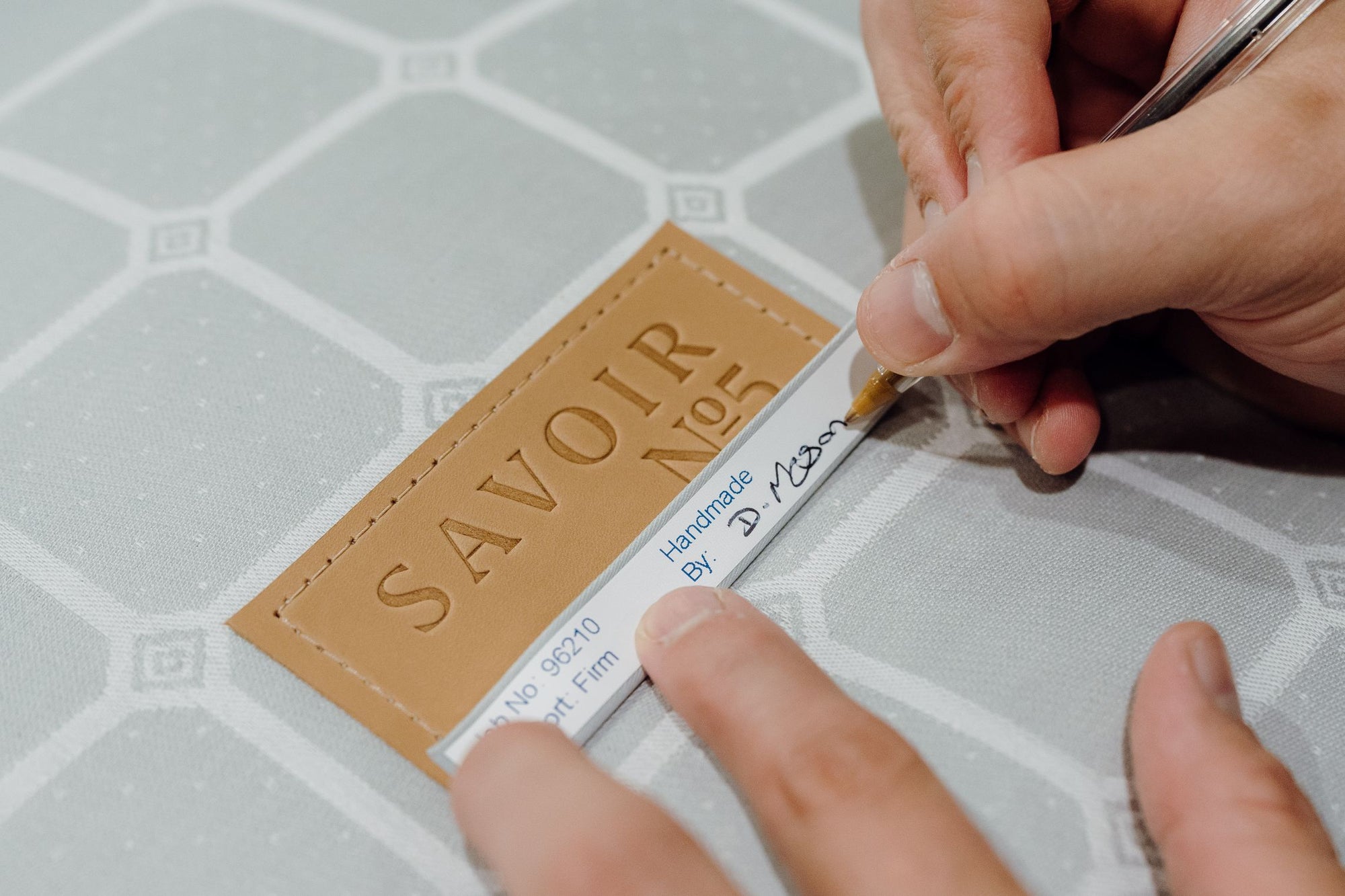 A close-up of a craftsman's hands writing his initials with a black pen on a label. The label features the embossed text 'SAVOIR No5'.