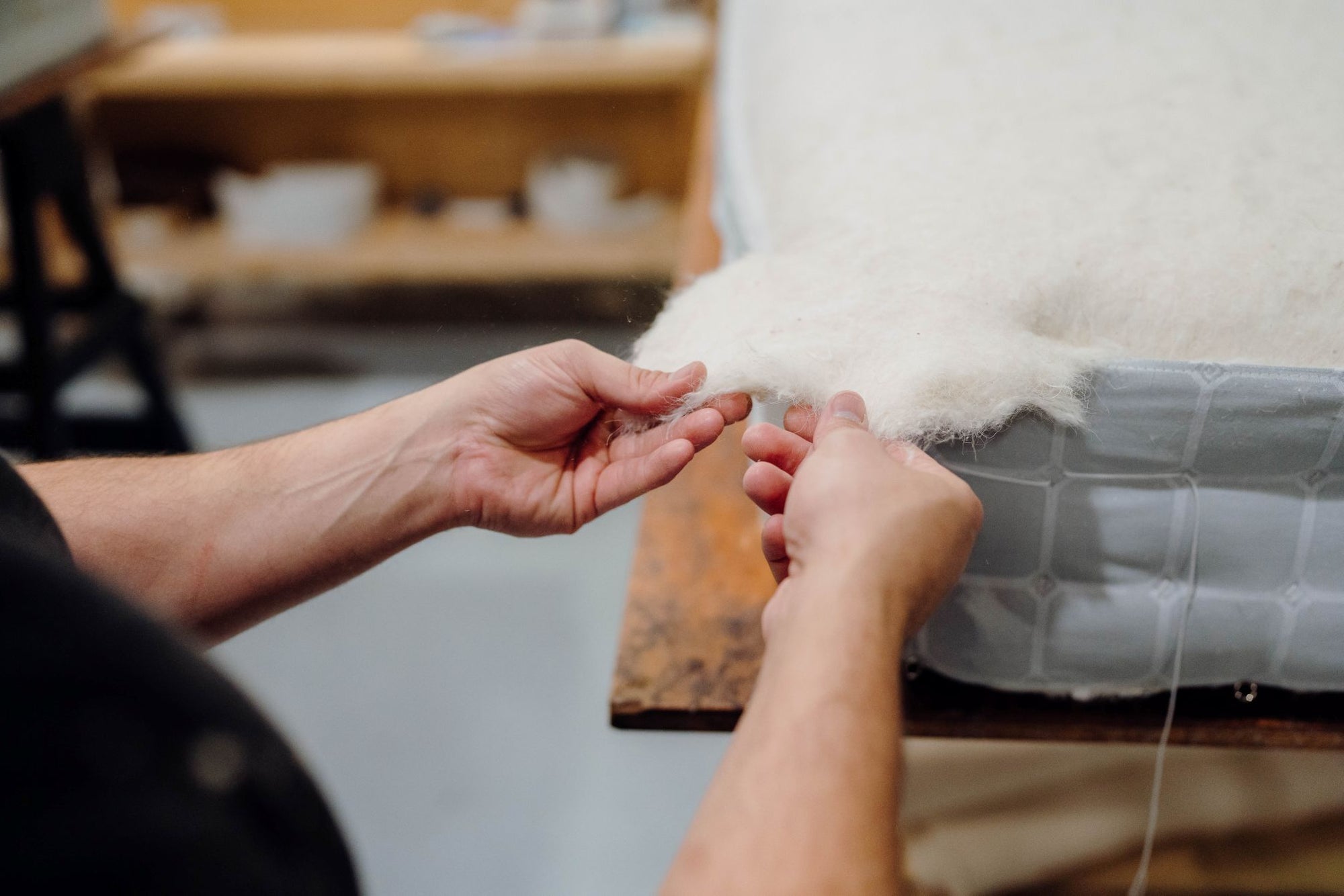 A close-up of a craftsman's hands adjusting wool to the edge of a Savoir mattress topper, set in the workshop.