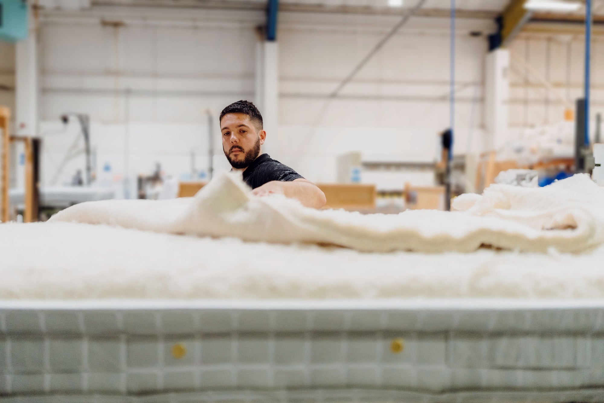 A craftsman in a black t-shirt sits behind a large mattress in a bright, spacious workshop setting with industrial equipment in the background.