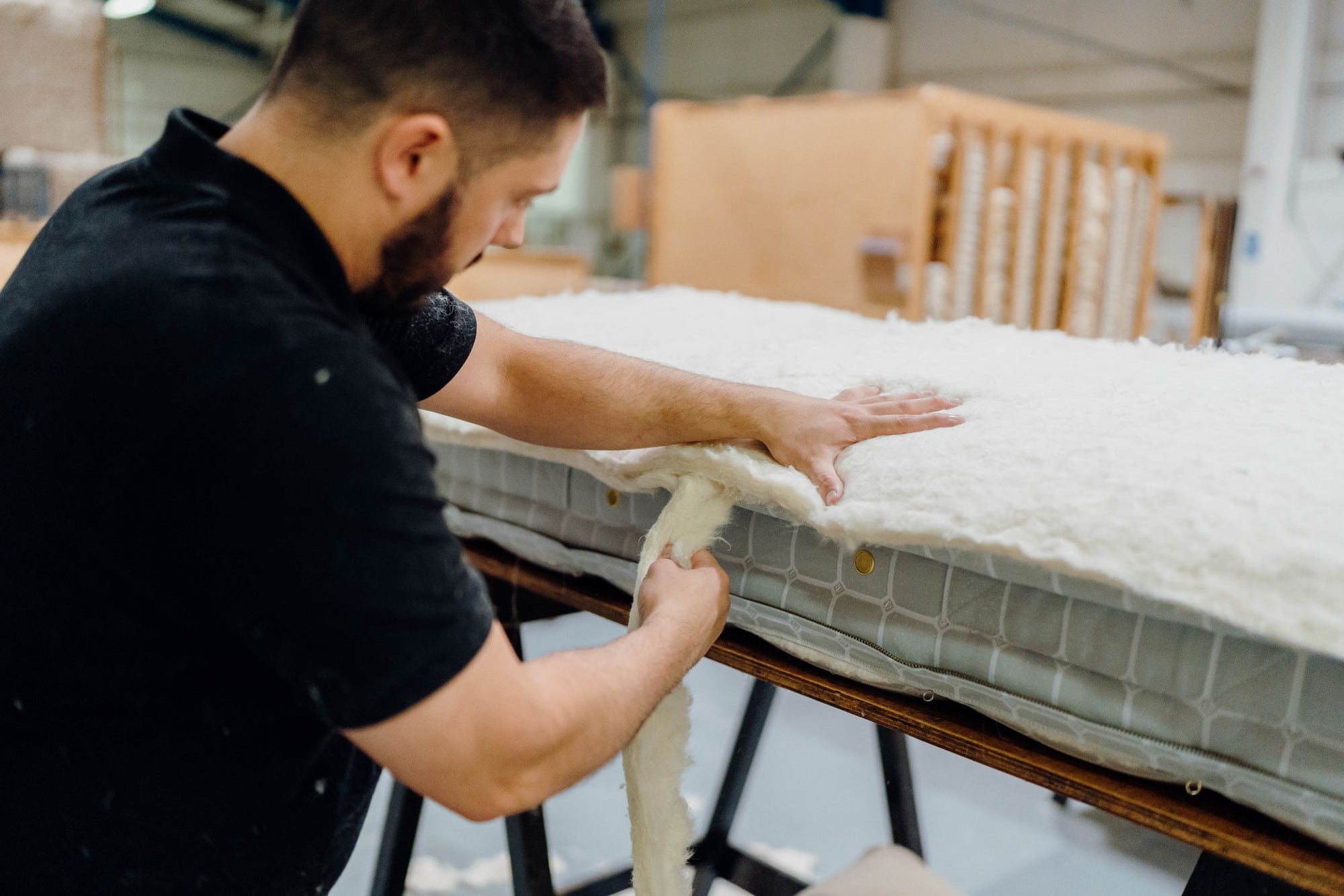 A Savoir craftsman in a black shirt works on a mattress in a workshop, peeling off a layer of wool from the side of the mattress frame.