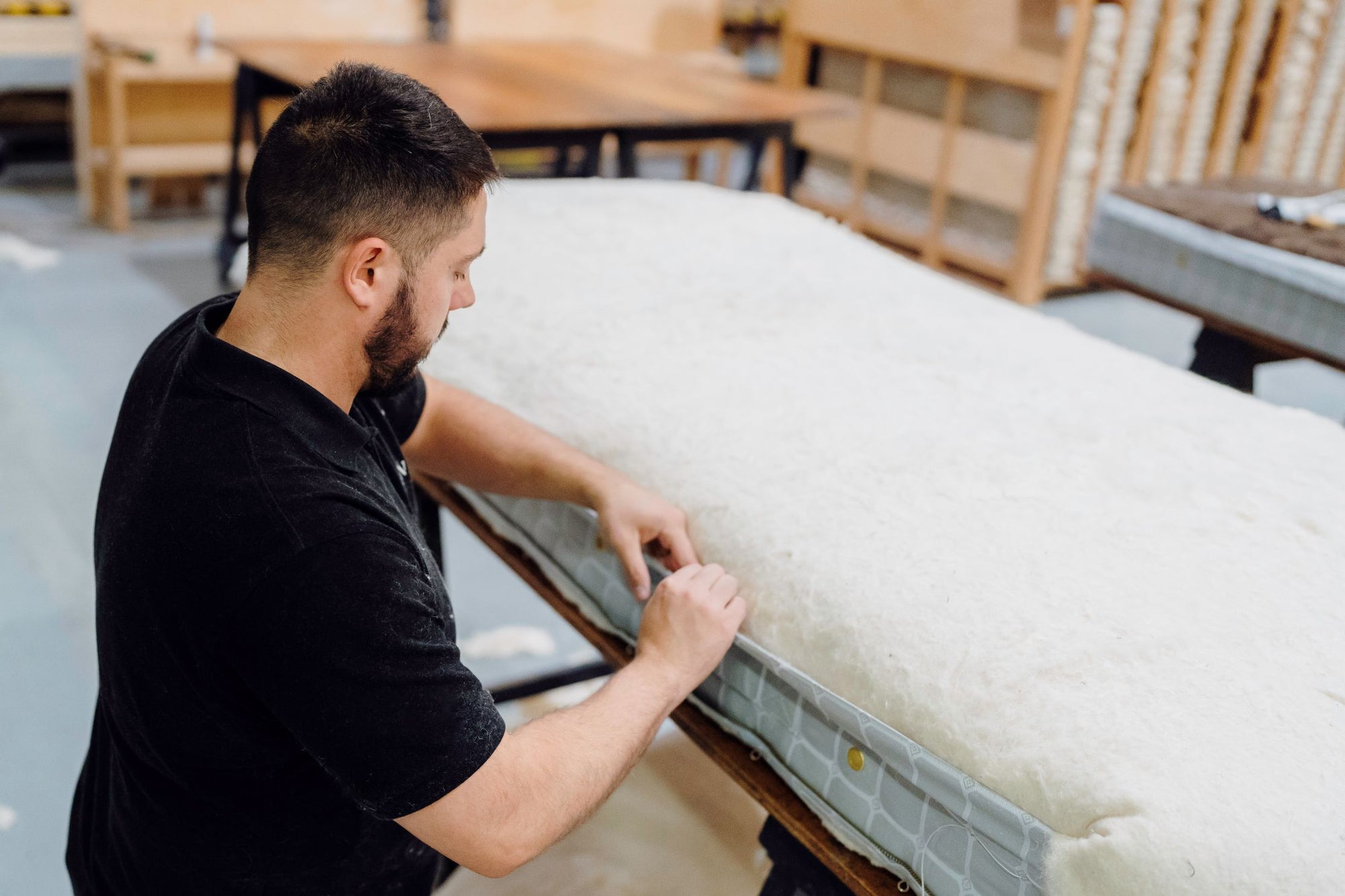 A craftsman in a black t-shirt carefully adjusting the side of a mattress on a wooden bed frame in the workshop.