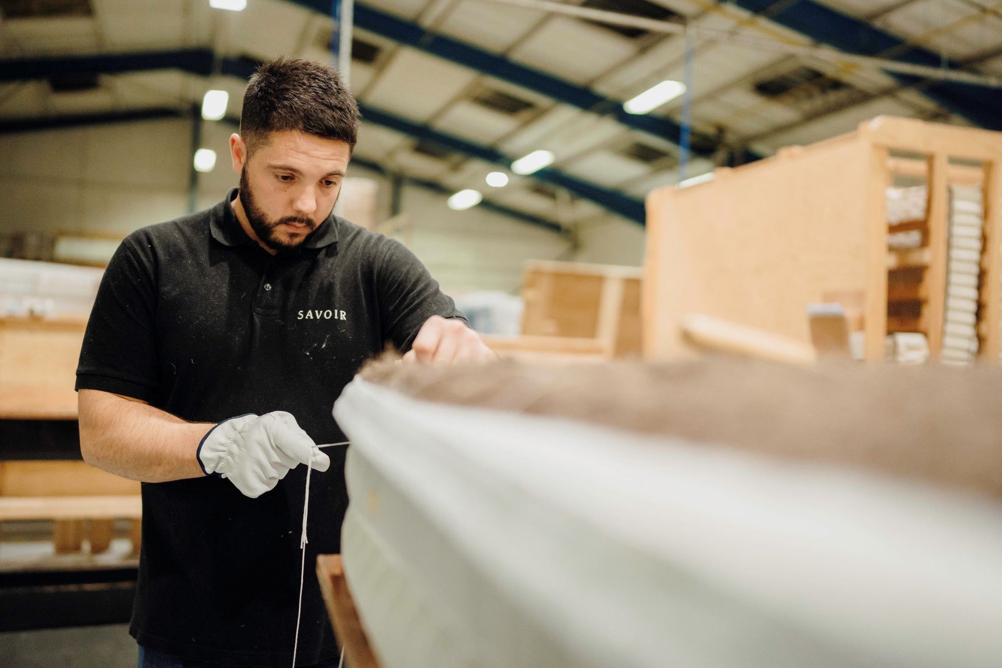 A craftsperson wearing a black Savoir polo shirt and white work gloves is seen in a workshop setting, holding a string near a mattress. The background shows wooden boat components and a high-ceilinged warehouse environment.