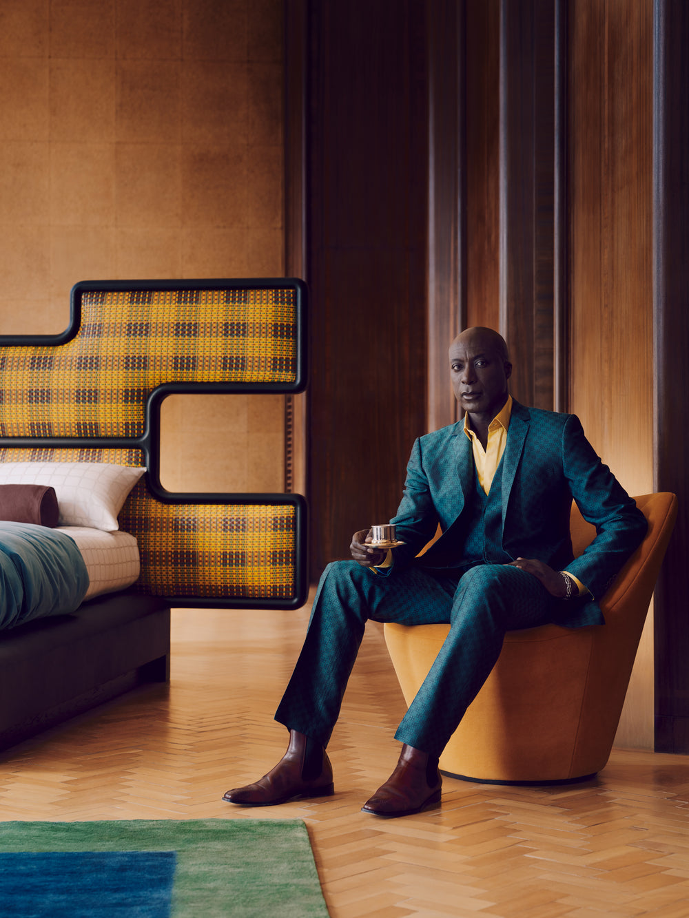 Fashion Designer Ozwald Boateng in a teal suit sitting on a modern chair in a stylish room with a bed and yellow and black patterned headboard.