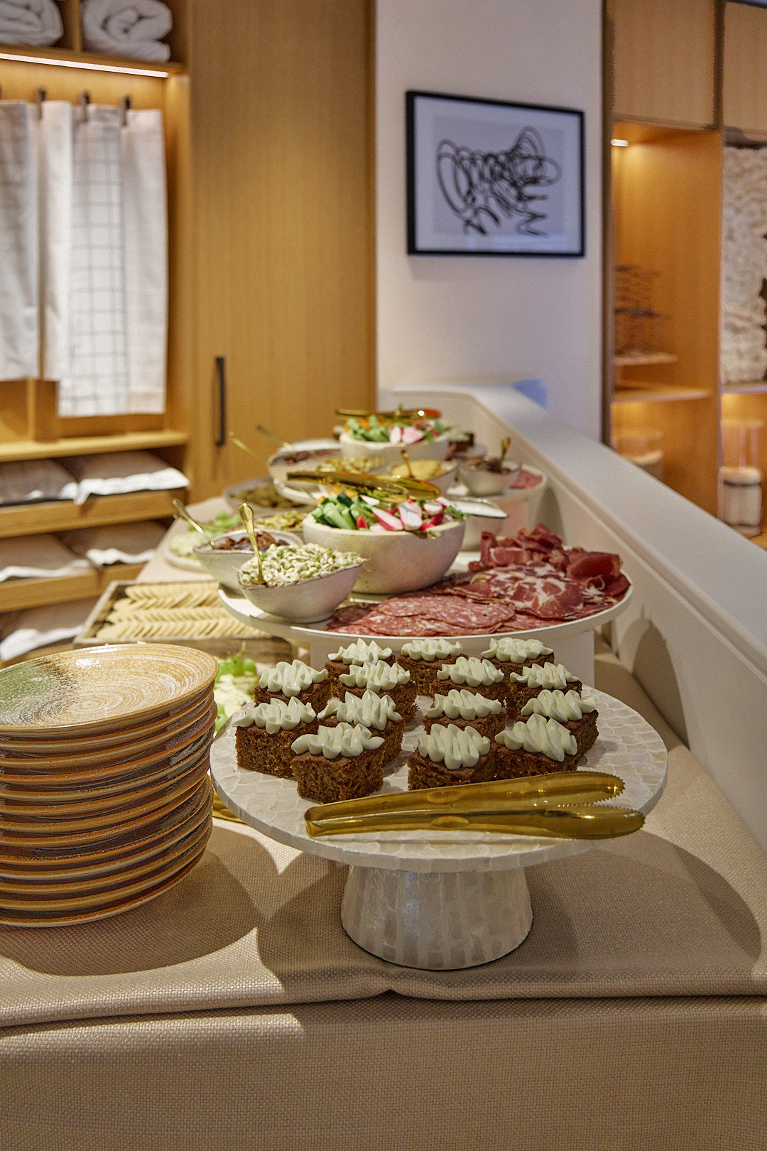 A buffet-style food spread on a table with plates, bowls of salad and sliced meat, and small dessert pieces, set in the Savoir Mayfair Showroom with shelves and framed wall art in the background.