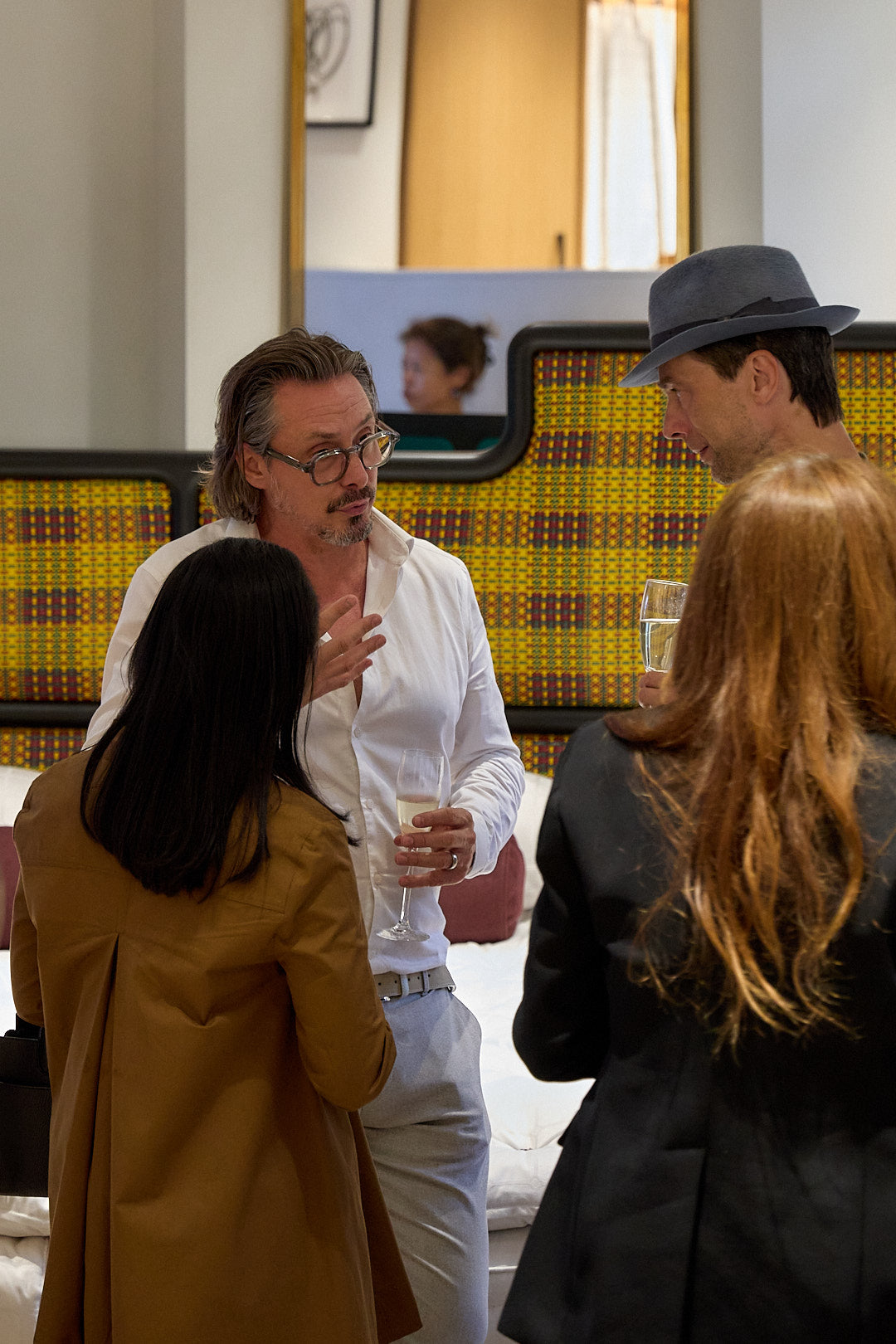 Three people conversing indoors in the Mayfair showroom with a yellow and brown patterned Authenticity headboard. One man wears a white shirt and light trousers, another wears a brown coat, and the third wears a black blazer and a dark fedora hat.