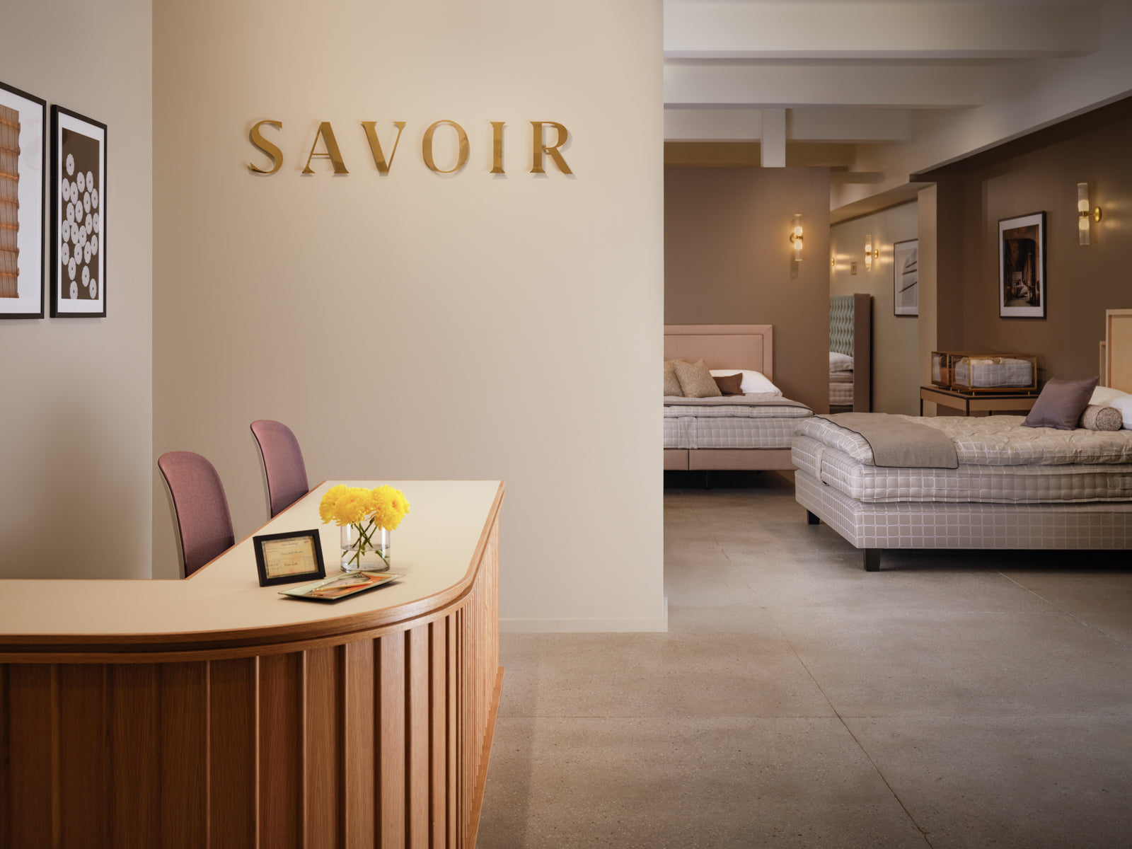 Our showroom interior located in New-York 200 Lex featuring a curved wooden reception desk in the foreground, a beige wall with gold 'SAVOIR' lettering, and multiple bed suites visible in the background.