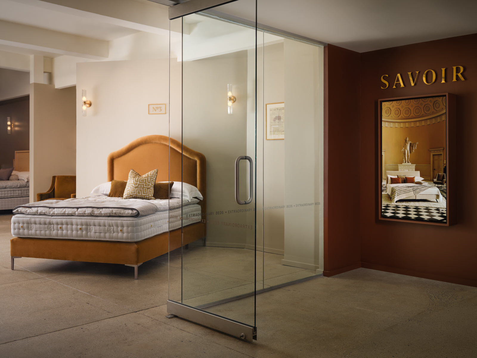 Savoir showroom featuring a glass sliding door, a bed with an orange headboard, topper and mattress, and a dark brown wall with gold 'SAVOIR' lettering and a framed Savoir bed picture.