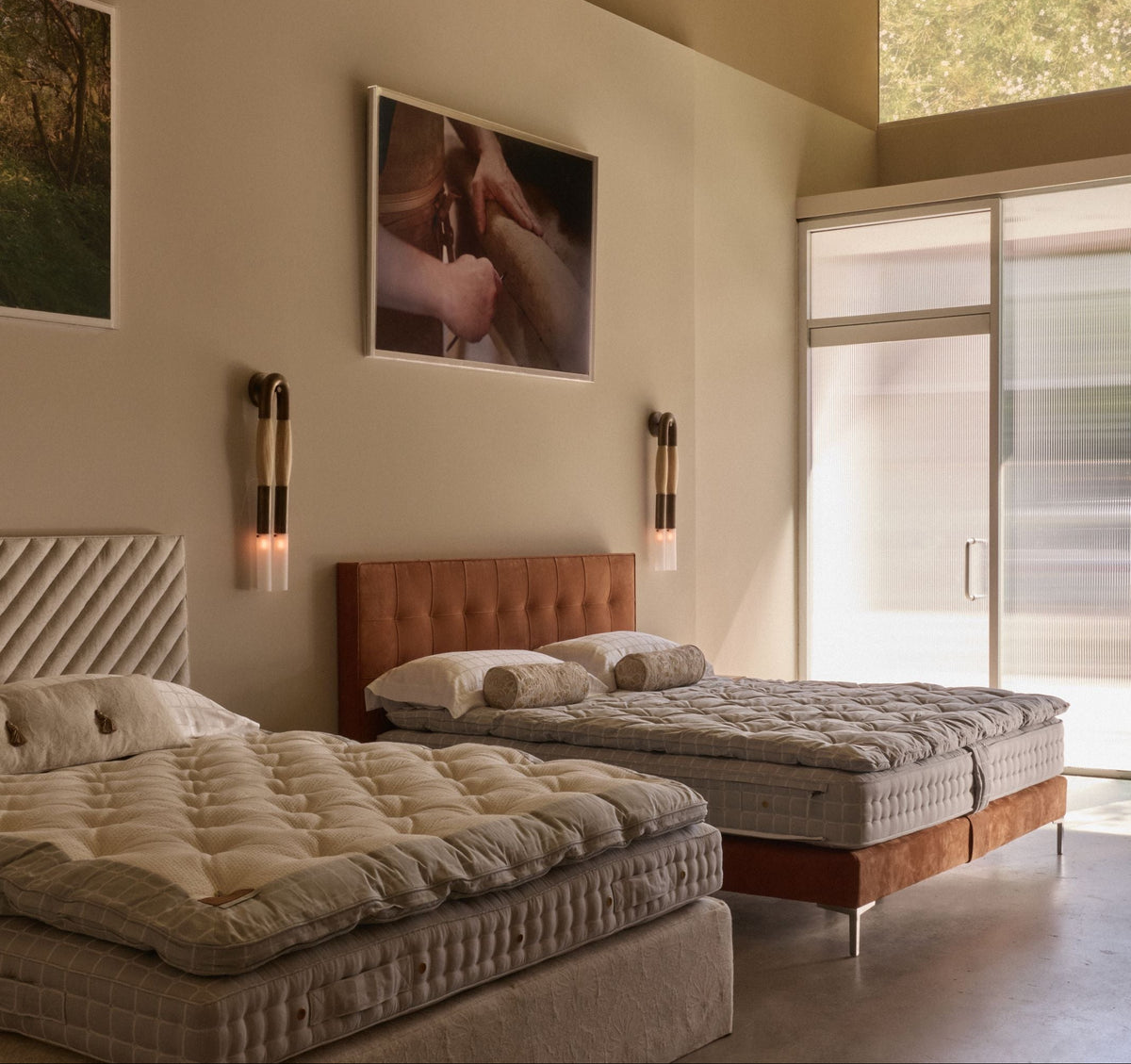The Los Angeles showroom interior featuring two beds with quilted beige bedding, a tufted brown headboard, wall-mounted sconces, framed wall art, and a large window.
