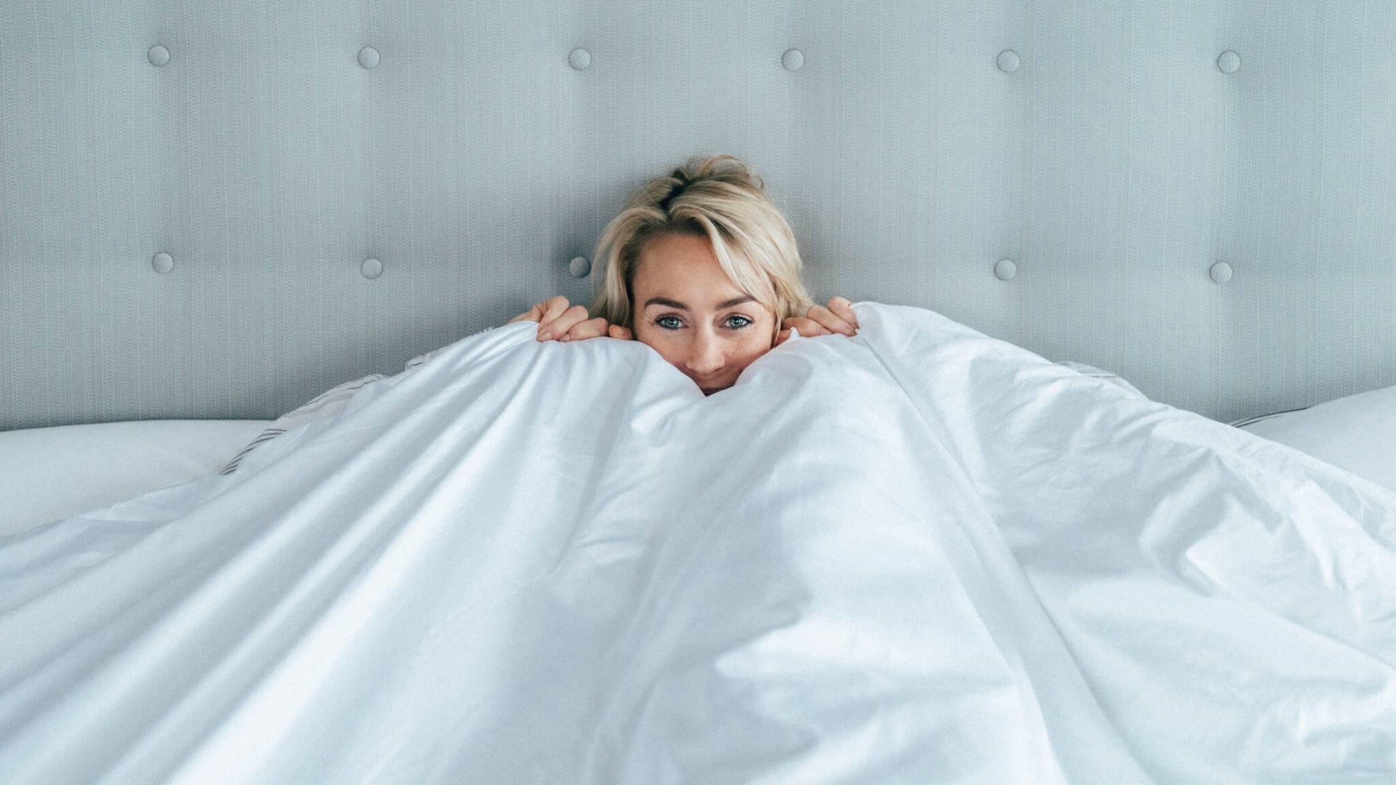 A blonde woman in a handmade king sized bed holding a duvet up to her face