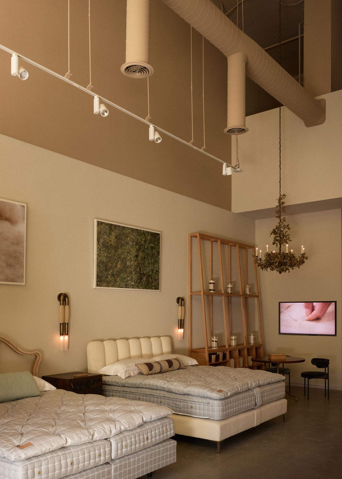The Savoir Los Angeles showroom featuring two beds, wall-mounted lamps, shelving, a hanging chandelier, and a television screen.