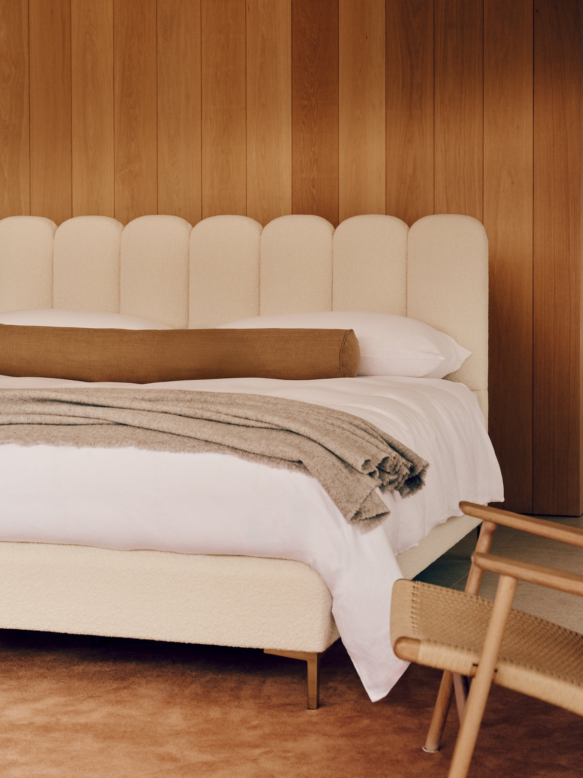 A modern upholstered bed with a cream scalloped headboard and matching footboard, styled with white bedding, a taupe cylindrical pillow, and a textured taupe throw blanket, set against a warm wood-paneled wall.