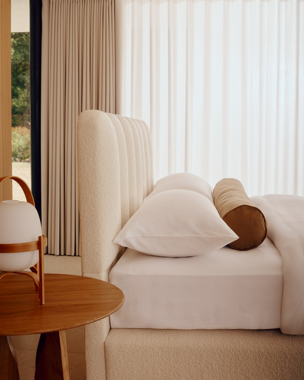 A modern bedroom featuring the textured cream bouclé Melrose bed with a tall channel-tufted headboard, styled with white pillows and a brown cylindrical bolster pillow. A matching cream bouclé base is visible at the bottom, and a round wooden side table with a white and wood lamp sits on the left.