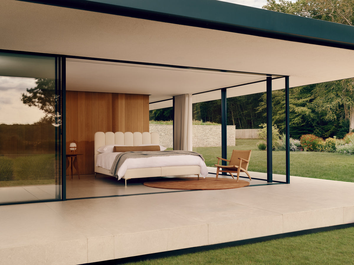 A modern bedroom interior with the Melrose bed, bedside table, chair, and large floor-to-ceiling glass walls, set within a contemporary house and overlooking a green lawn.