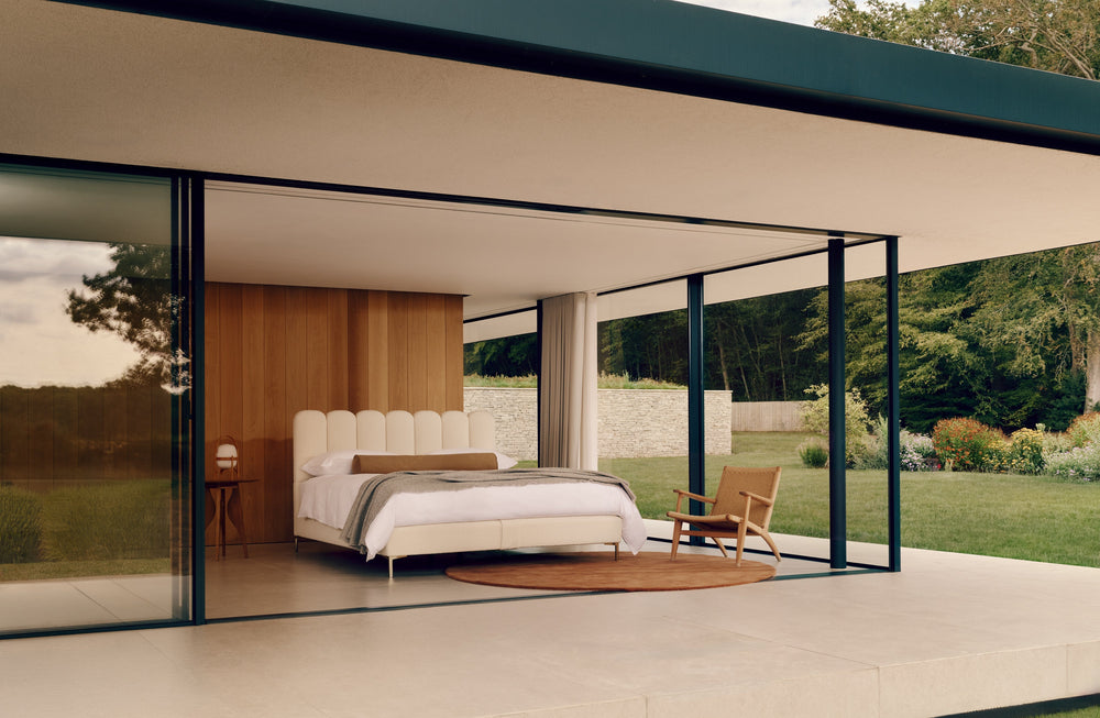 The Melrose bed with in a beautiful setting white bedding, a tufted headboard, and an open-plan design with floor-to-ceiling glass walls, overlooking a landscaped garden and patio.