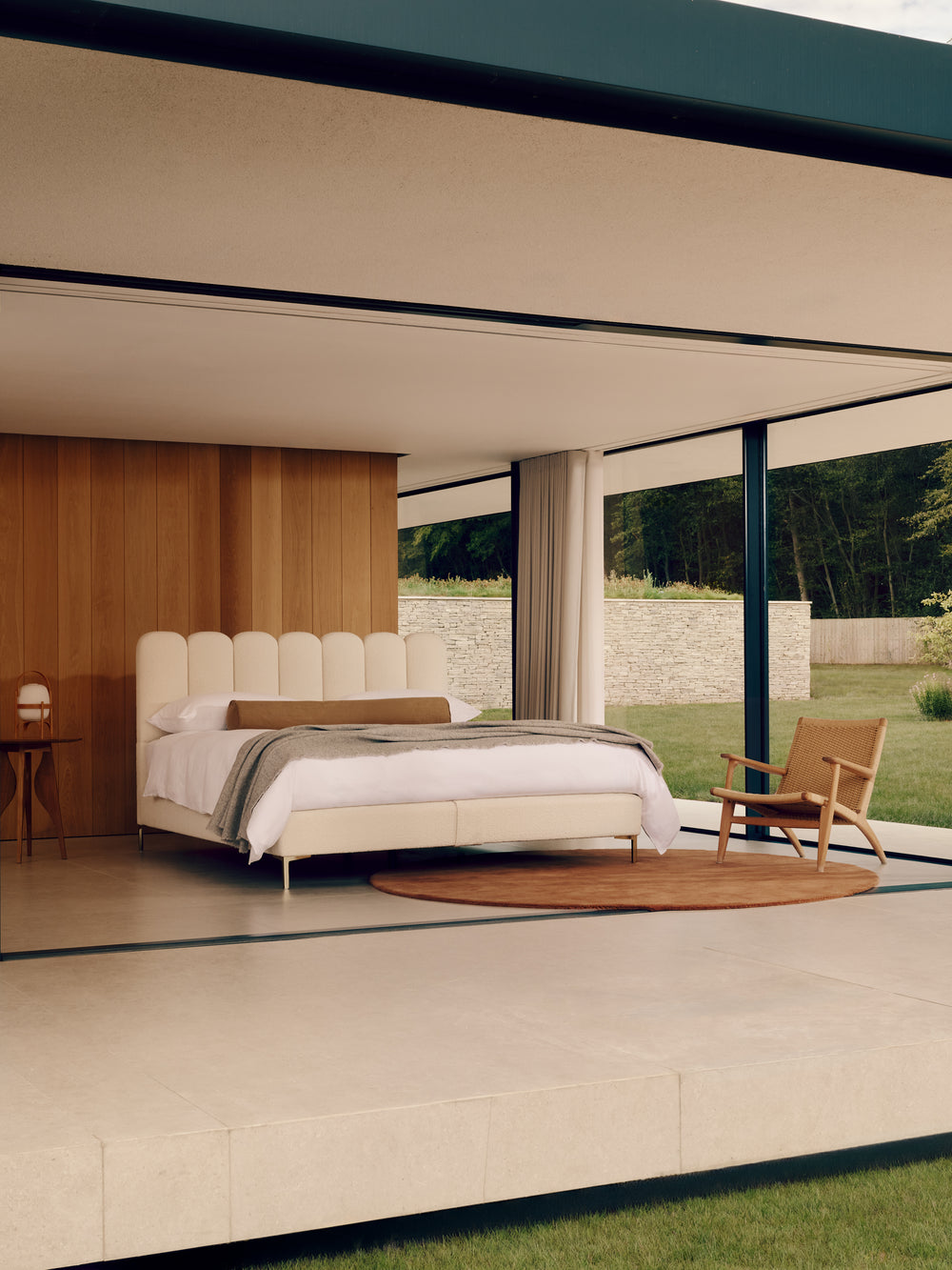 A modern bedroom interior with the Melrose bed dressed in white bedding, a tufted headboard, a round brown rug, a wooden chair, and large floor-to-ceiling windows overlooking a garden.