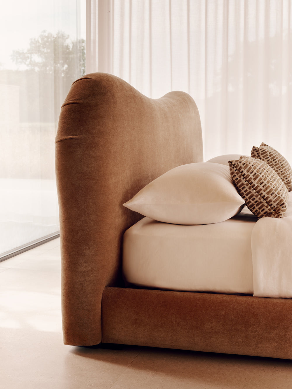 The Teddy bed with a curved upholstered headboard in warm camel brown velvet, styled with cream bedding and two textured brown and beige pillows in a bright, minimalist bedroom.