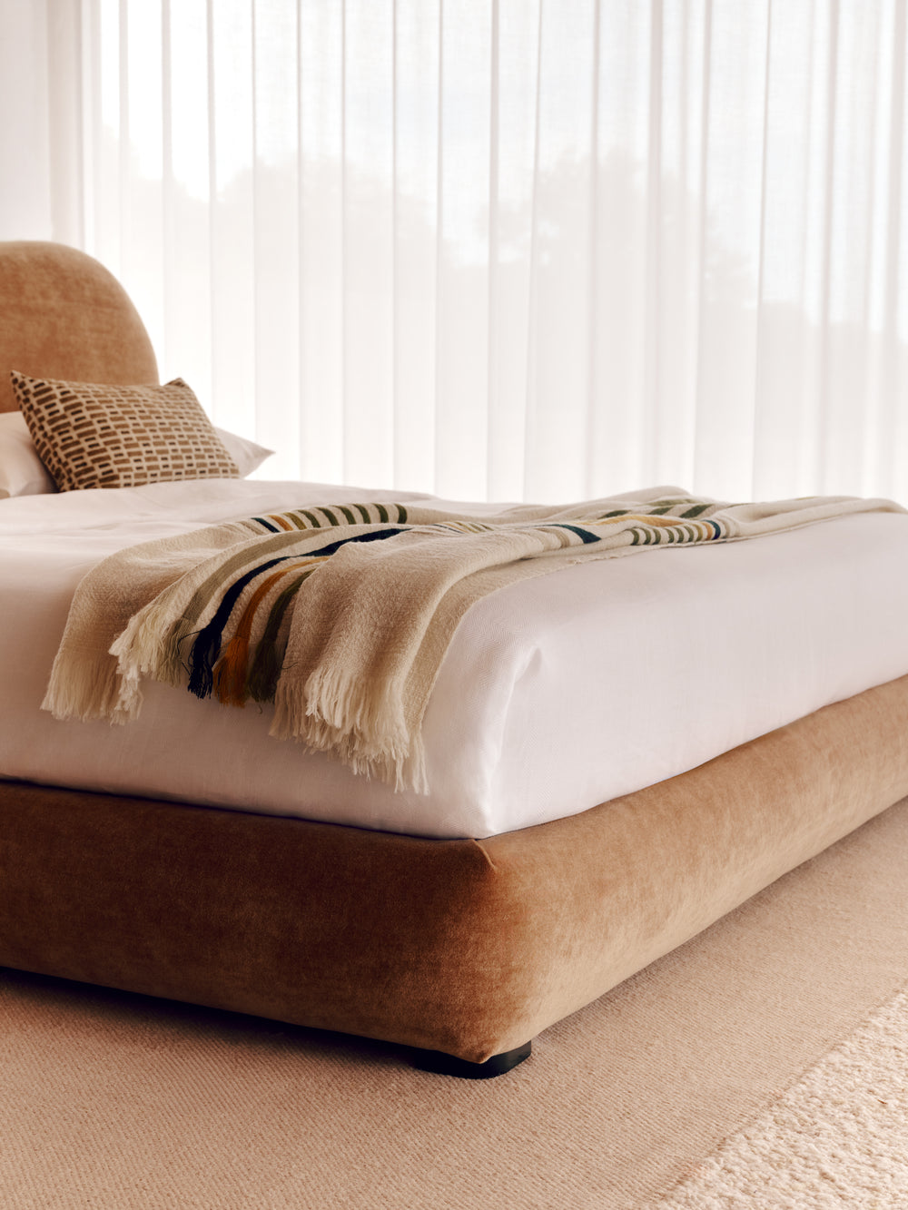The low-profile upholstered Teddy bed with a warm taupe velvet frame, dressed in white bedding and a striped beige throw blanket, set in a bright bedroom with sheer curtains.