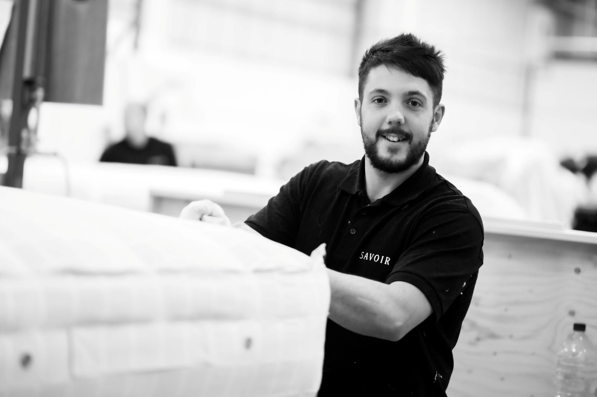 A black and white photo of a smiling craftsman with a beard wearing a black polo shirt with the brand name 'SAVOIR' on the chest, holding a mattress in the workshop.