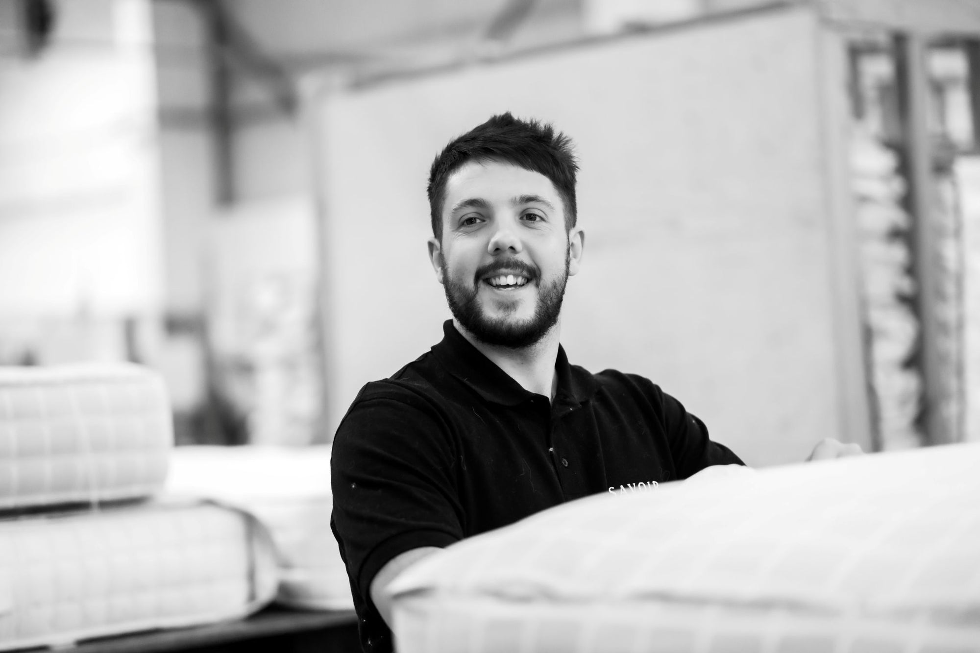 A black and white portrait of a craftsman with a beard wearing a dark polo shirt, standing in a bedding  workshop with upholstered mattresses in the blurred background.