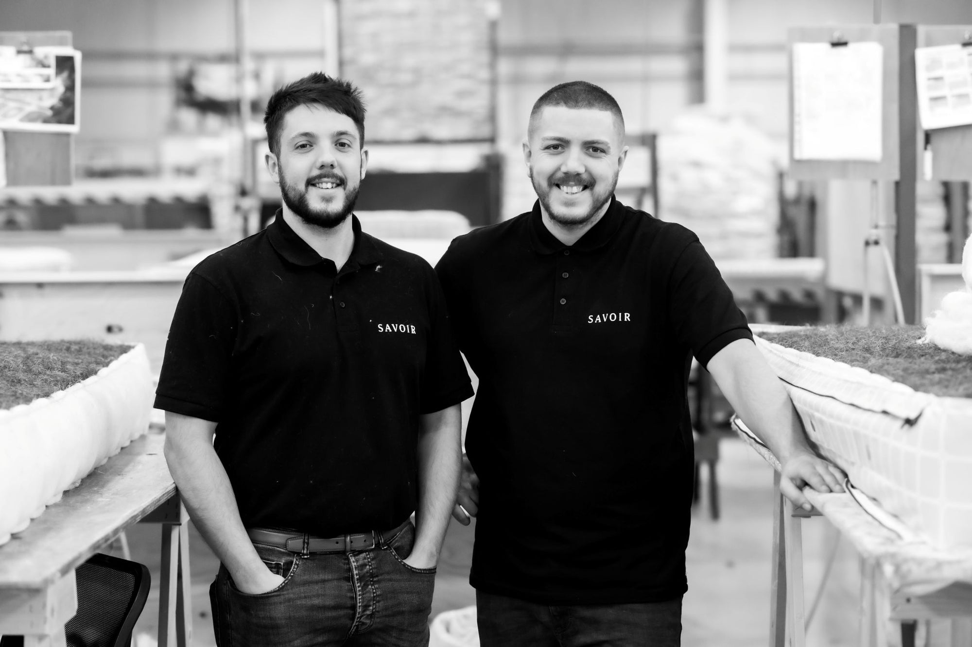 Two craftsmen standing side-by-side in a black polo shirt with the brand name 'SAVOIR' printed on the chest, in the workshop.