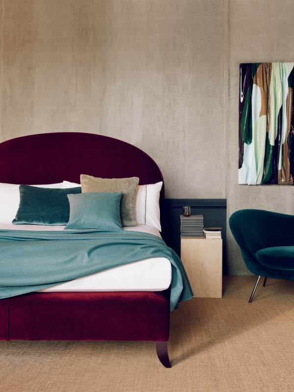 Image of the Elly design, photographed against brutalist concrete walls. The bed is upholstered in rich ruby velvet and dressed in luxurious white bed linen and ocean-coloured throws and cushions.