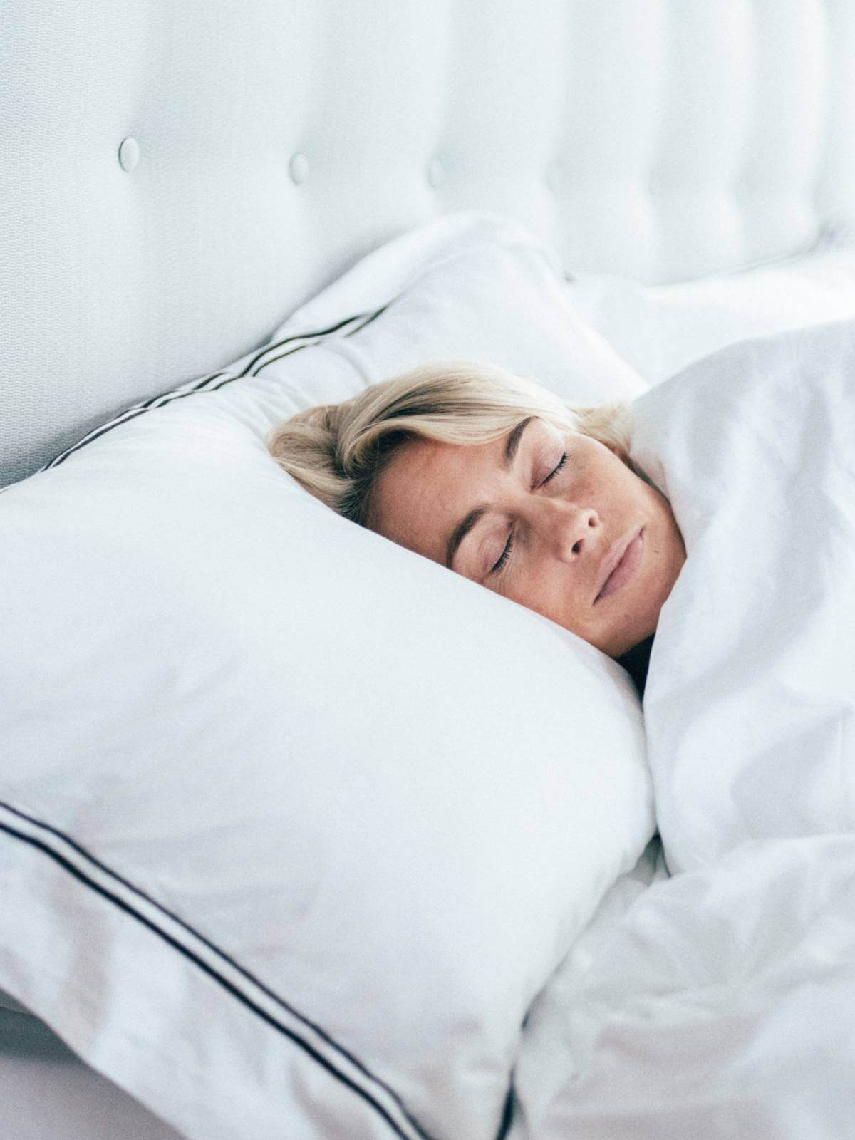 Woman sleeping on her side on a plush pillow, dressed in all-white bed linen with charcoal cording.