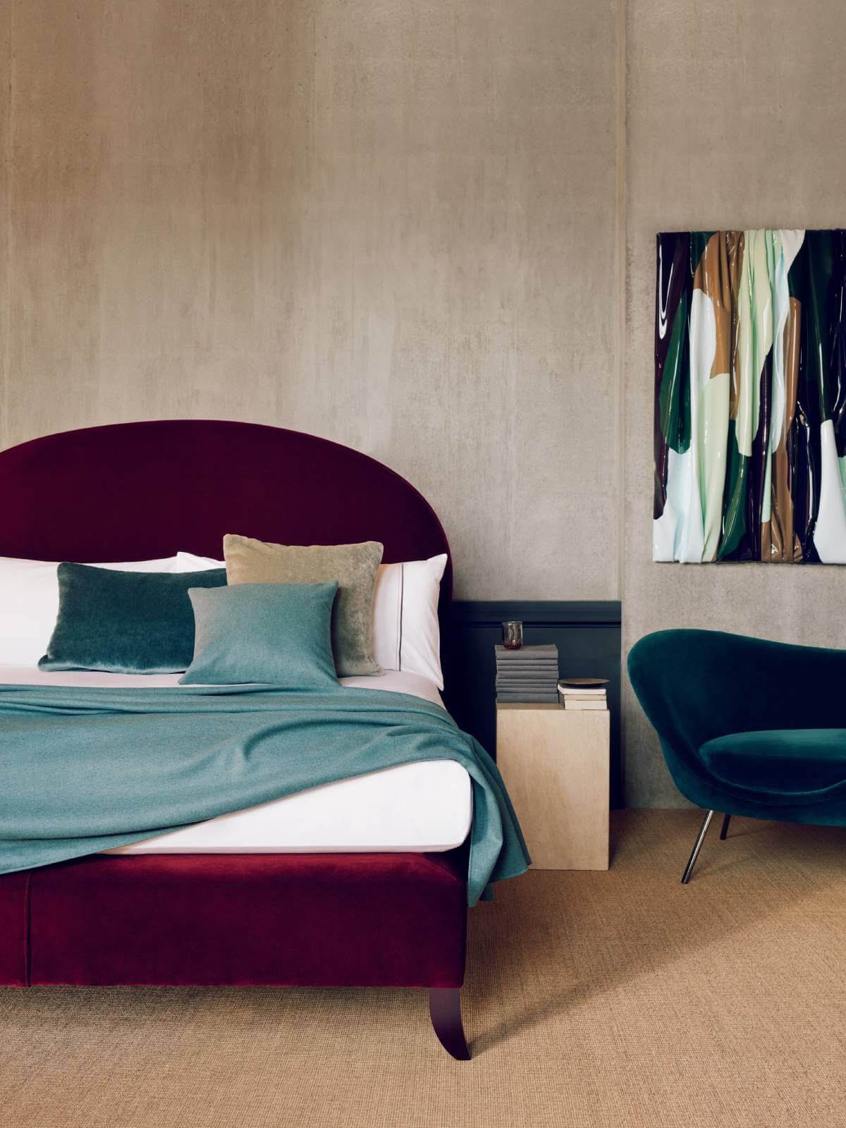 Image of the Elly design, photographed against brutalist concrete walls. The bed is upholstered in rich ruby velvet and dressed in luxurious white bed linen and ocean-coloured throws and cushions.