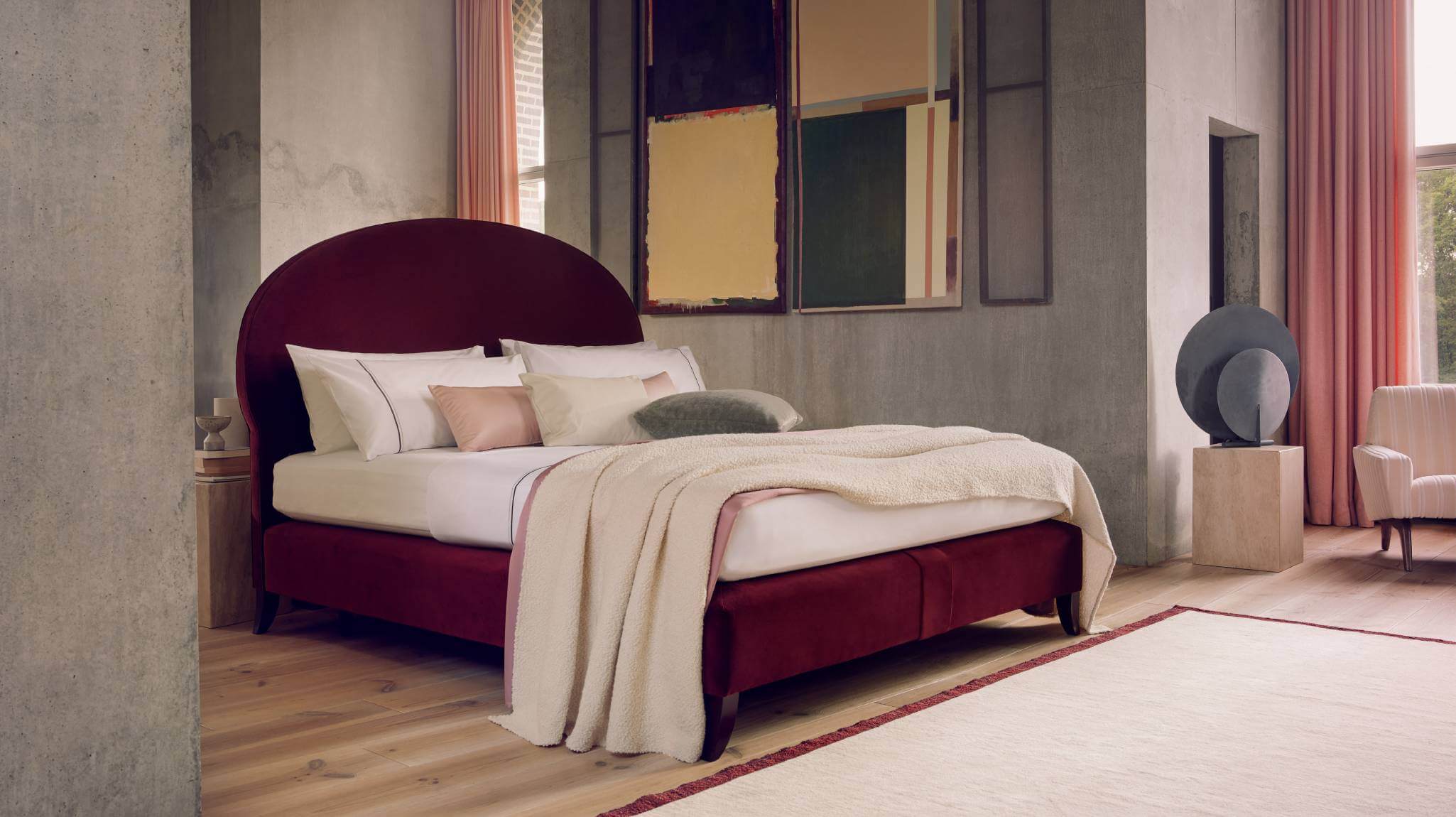 Image of the Elly design, photographed against brutalist concrete walls. The bed is upholstered in rich ruby velvet and dressed in luxurious white bed linen and ocean-coloured throws and cushions.