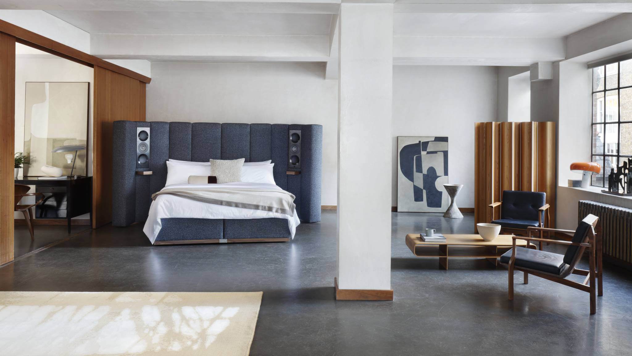 Image of our Seventy Five bed, featuring a pair of speakers integrated onto curved panels upholstered in textured blue fabric, photographed against mid-century-modern furniture and limewashed walls.